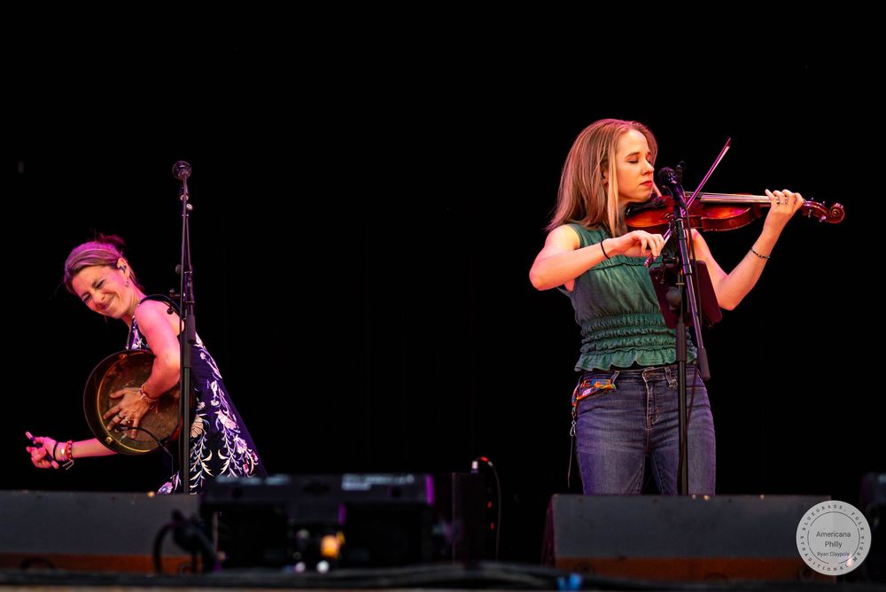 Two women perform on stage, one playing violin and one playing banjo, at an outdoor music festival.