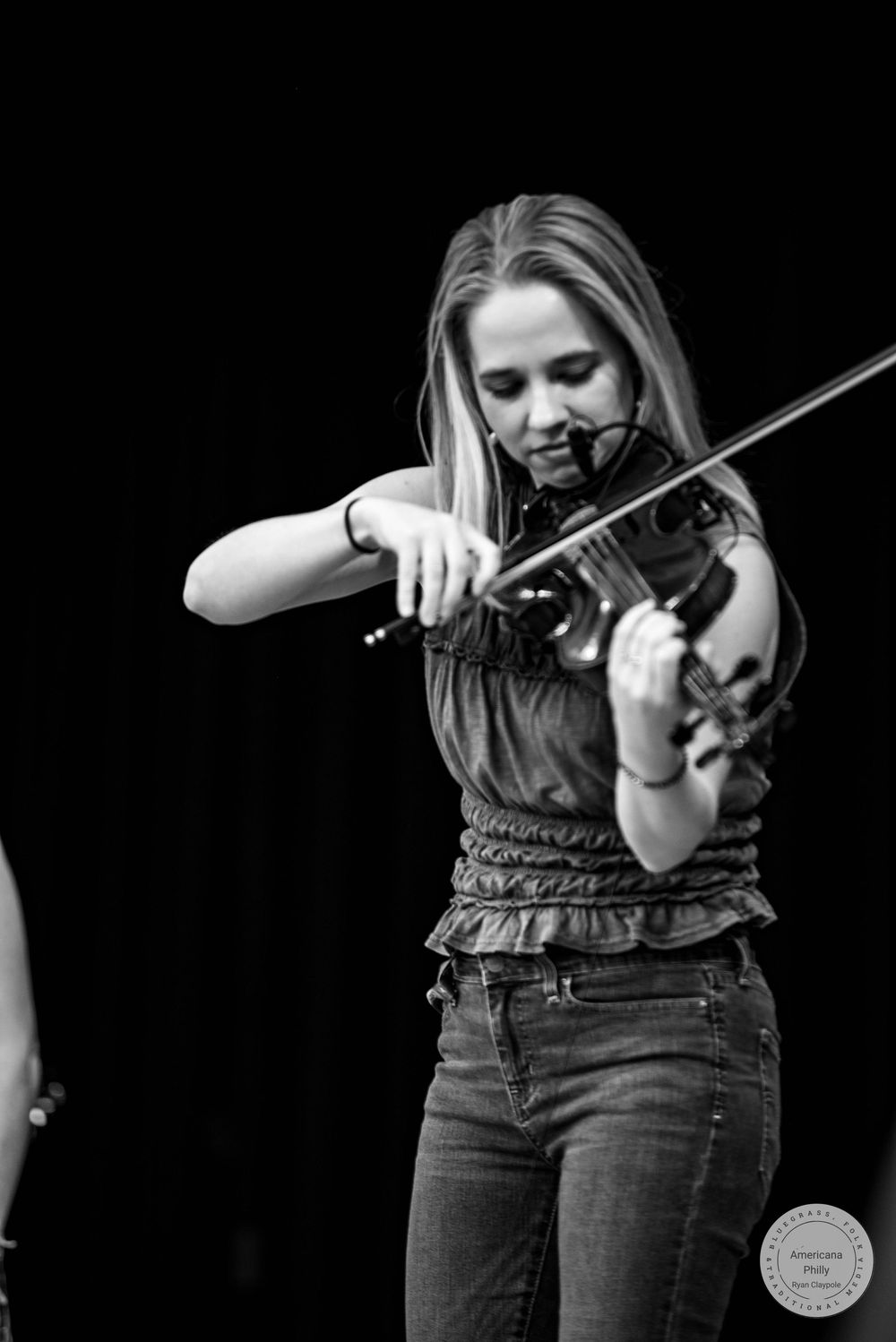 Black and white photo of a young woman playing violin on stage, wearing a sleeveless top and jeans.