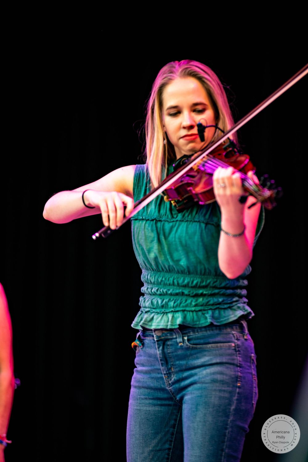 Young woman in green top plays violin on stage under pink spotlight, performing live music concert.