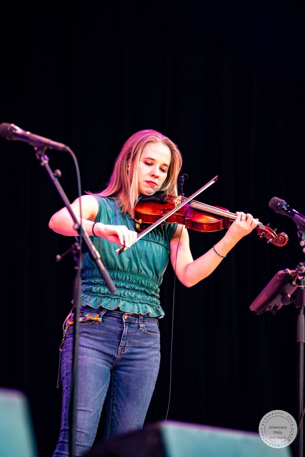 Young woman in green top and jeans playing violin on stage under pink stage lighting.