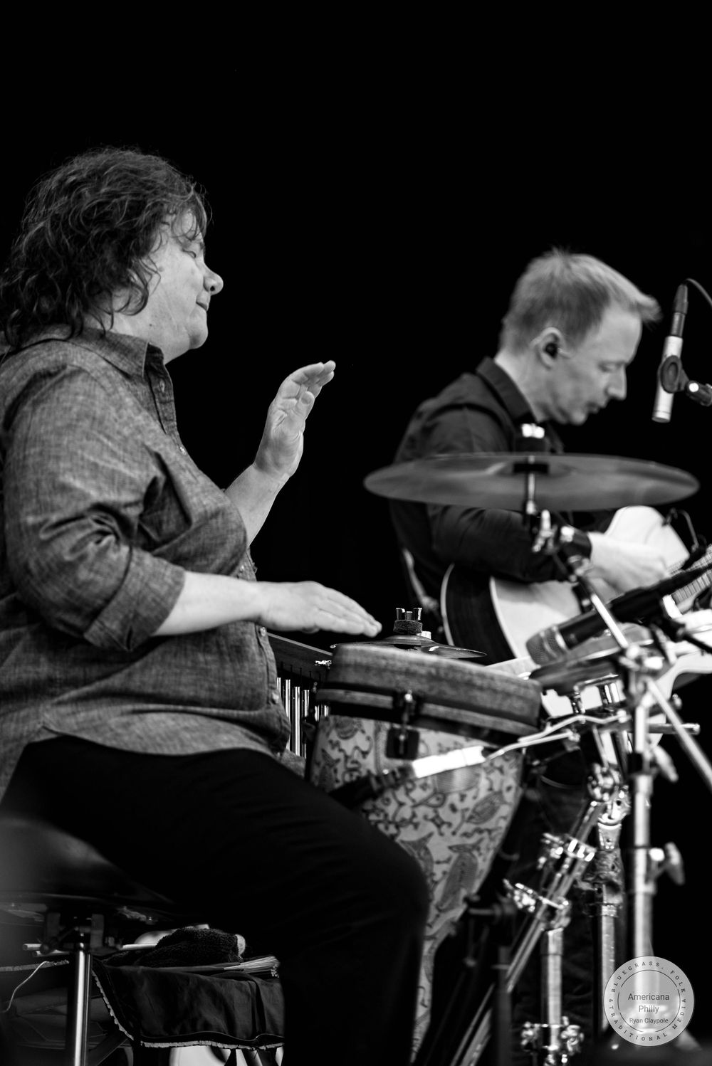 Black and white photo of two musicians performing live, one playing percussion and another on drums with cymbals visible.