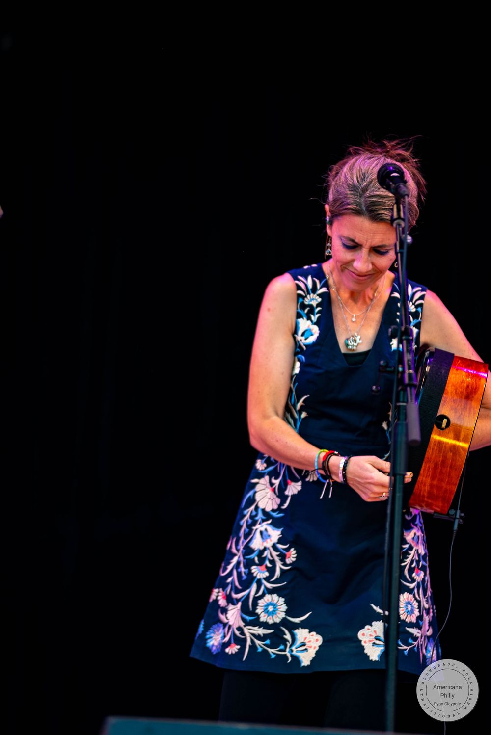 Woman in floral navy dress plays instrument on stage under warm spotlight at live music performance.