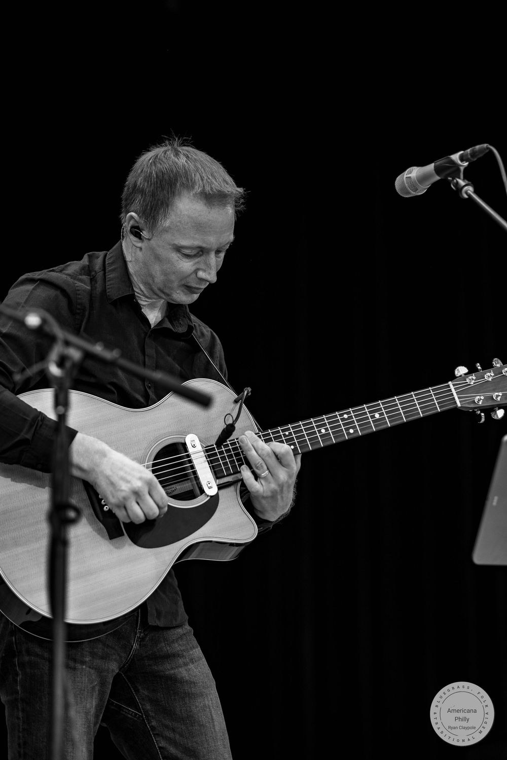 Black and white photo of a male guitarist playing acoustic guitar on stage with a microphone stand nearby.