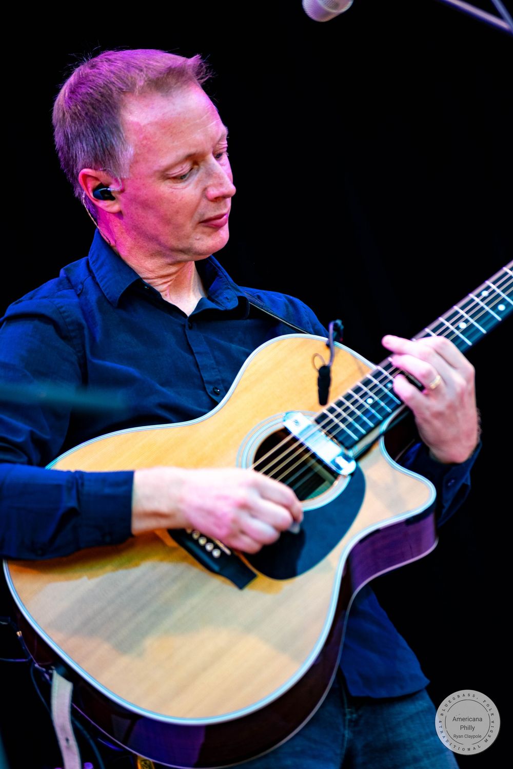 Man in dark shirt playing acoustic guitar on stage under purple stage lighting.
