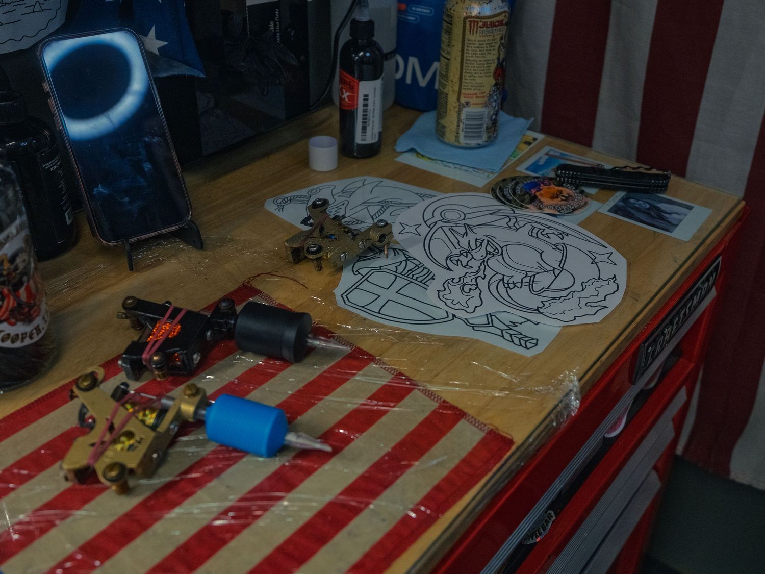 Tattoo machines and line-drawing stencils laid out on a flag-draped station at Sacred Art Tattoo in Honolulu, Hawaii, ink and phone nearby.