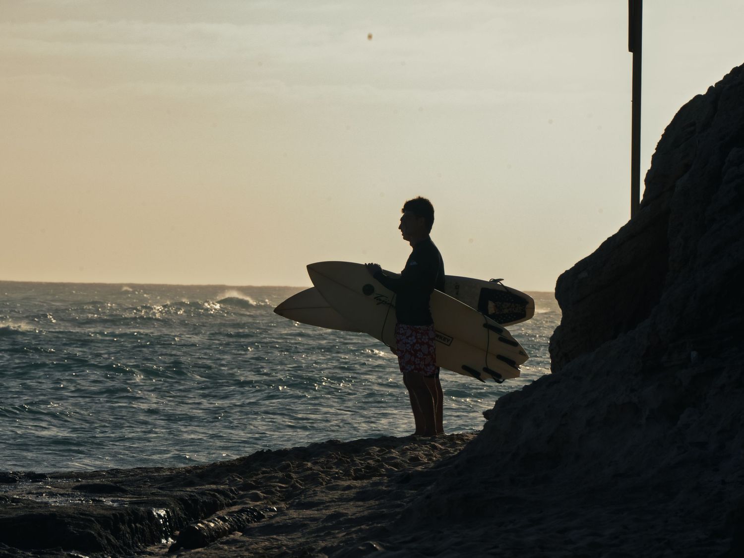 A surfer holding a pale surfboard stands on a rocky shoreline at Waikiki, silhouetted against a hazy golden sky, watching the waves.