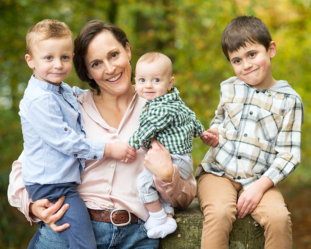 Smiling mother sits outdoors with three children: a toddler on her lap, a baby in green gingham, and an older boy beside her.