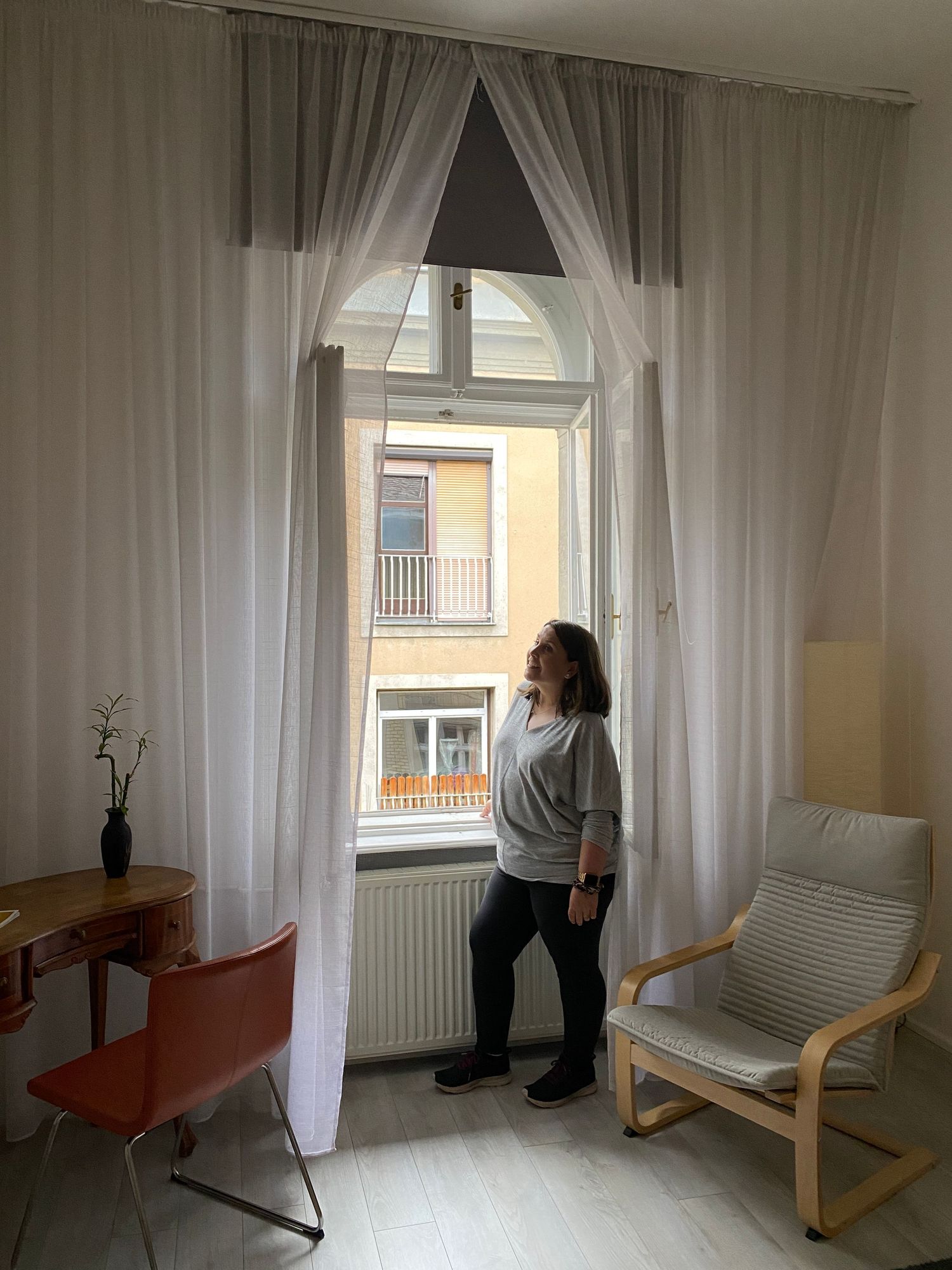 Woman standing by tall window with white curtains in a bright, minimalist room with wooden desk and modern chair.
