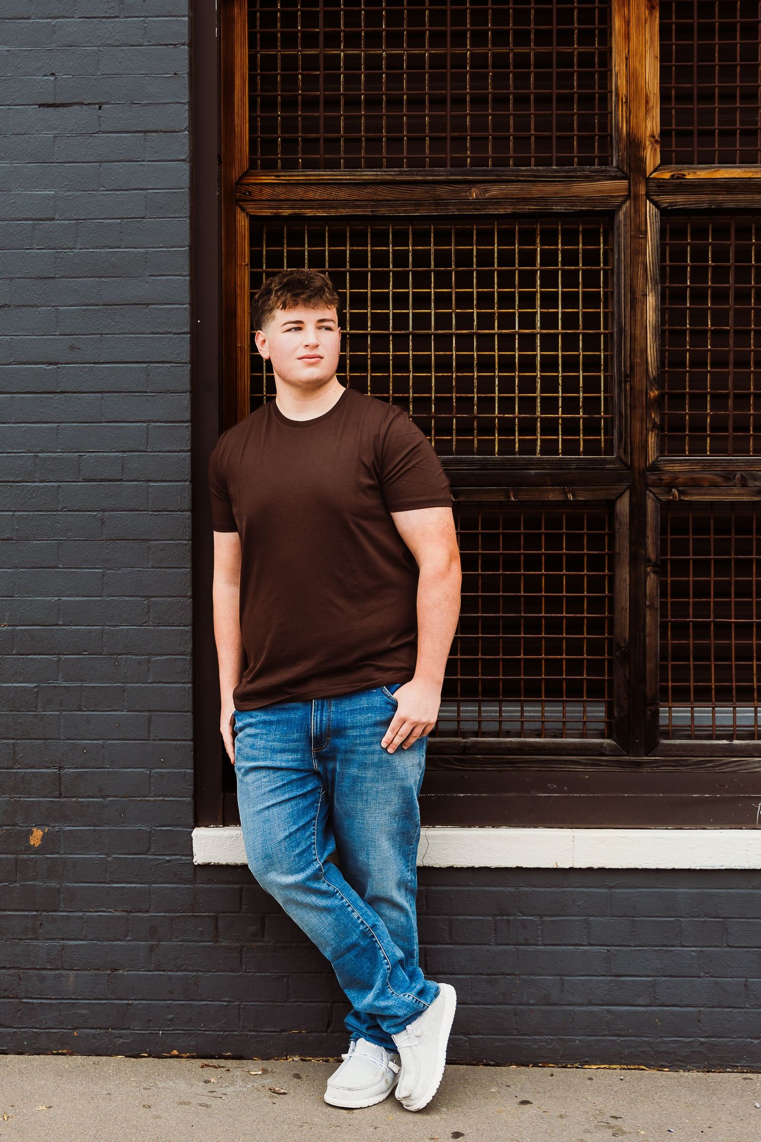 Young man in brown t-shirt and jeans leaning against gray brick wall with metal grid window.