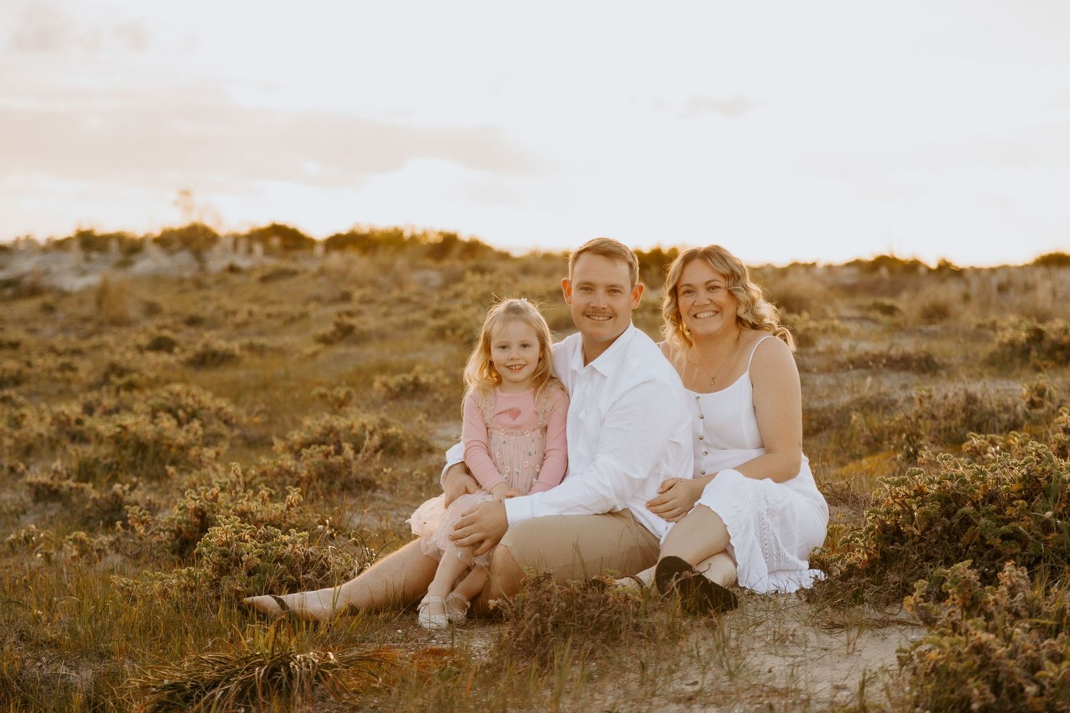 A smiling family of three sits in a golden field, captured by Emma Marie Photography at sunset.