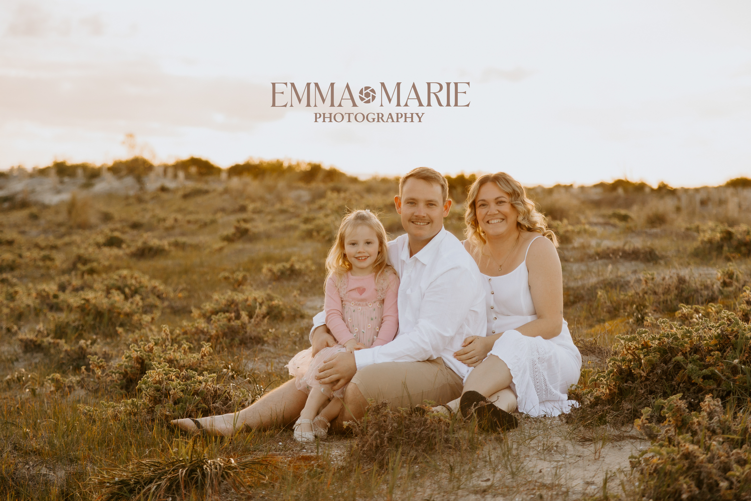 A smiling family of three sits in a golden field, captured by Emma Marie Photography at sunset.