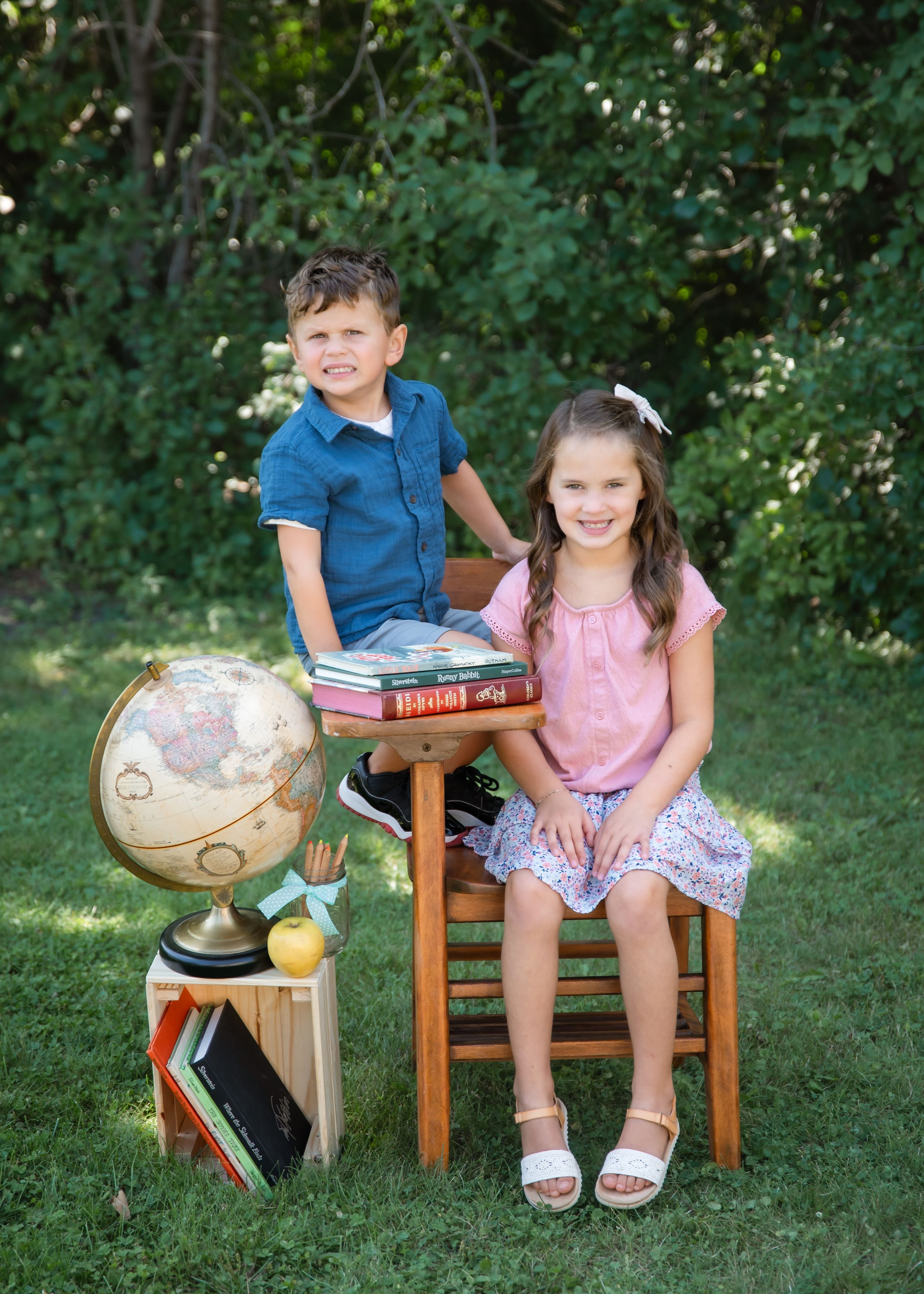 Two children pose outdoors with a globe and books for a back-to-school photo on green grass.