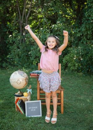 Excited girl in pink top and floral shorts sits on chair with arms raised, globe and 1st grade sign nearby.