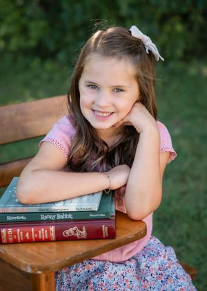 Young girl in pink shirt smiling on wooden bench with stack of books outdoors.