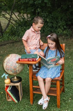 Two children reading books outdoors near a globe and stacked books on a small wooden stand in a garden setting.