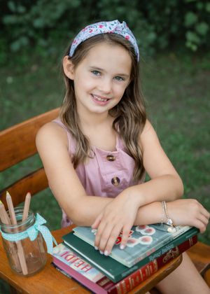 Young girl with floral headband smiles at desk with books and pencils outdoors.