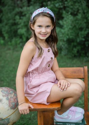 Young girl in pink dress and floral headband sitting on wooden chair with globe, smiling outdoors.
