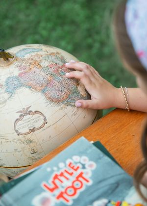 A child's hand points to a location on a vintage globe beside a Little Toot book on a wooden table.