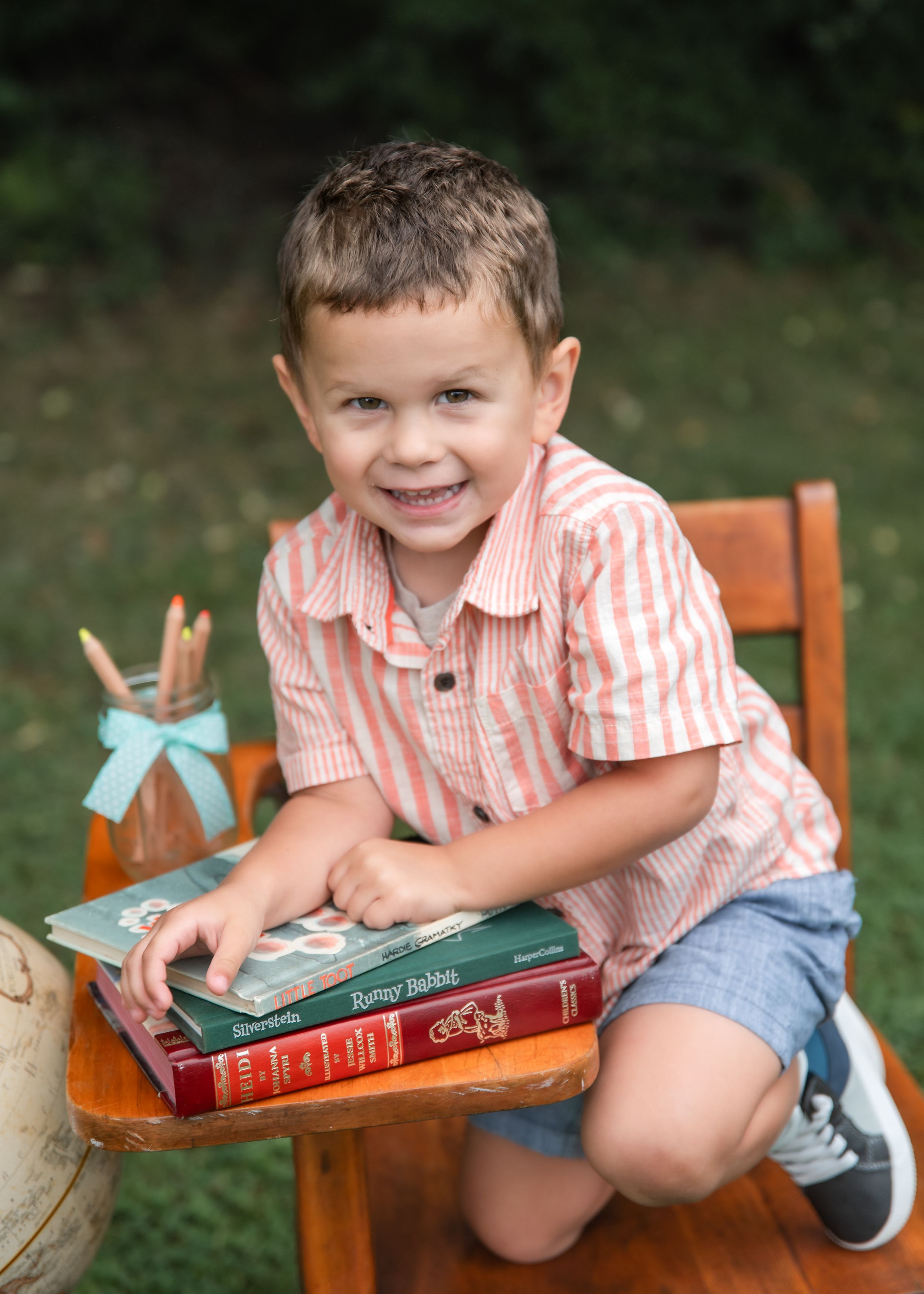 Smiling young boy in pink striped shirt kneeling on wooden chair with stacked books outdoors.