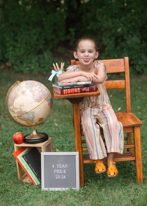 Young girl in striped jumpsuit sits on wooden chair with globe, books, and Pre-K 4 year sign for back to school photo.