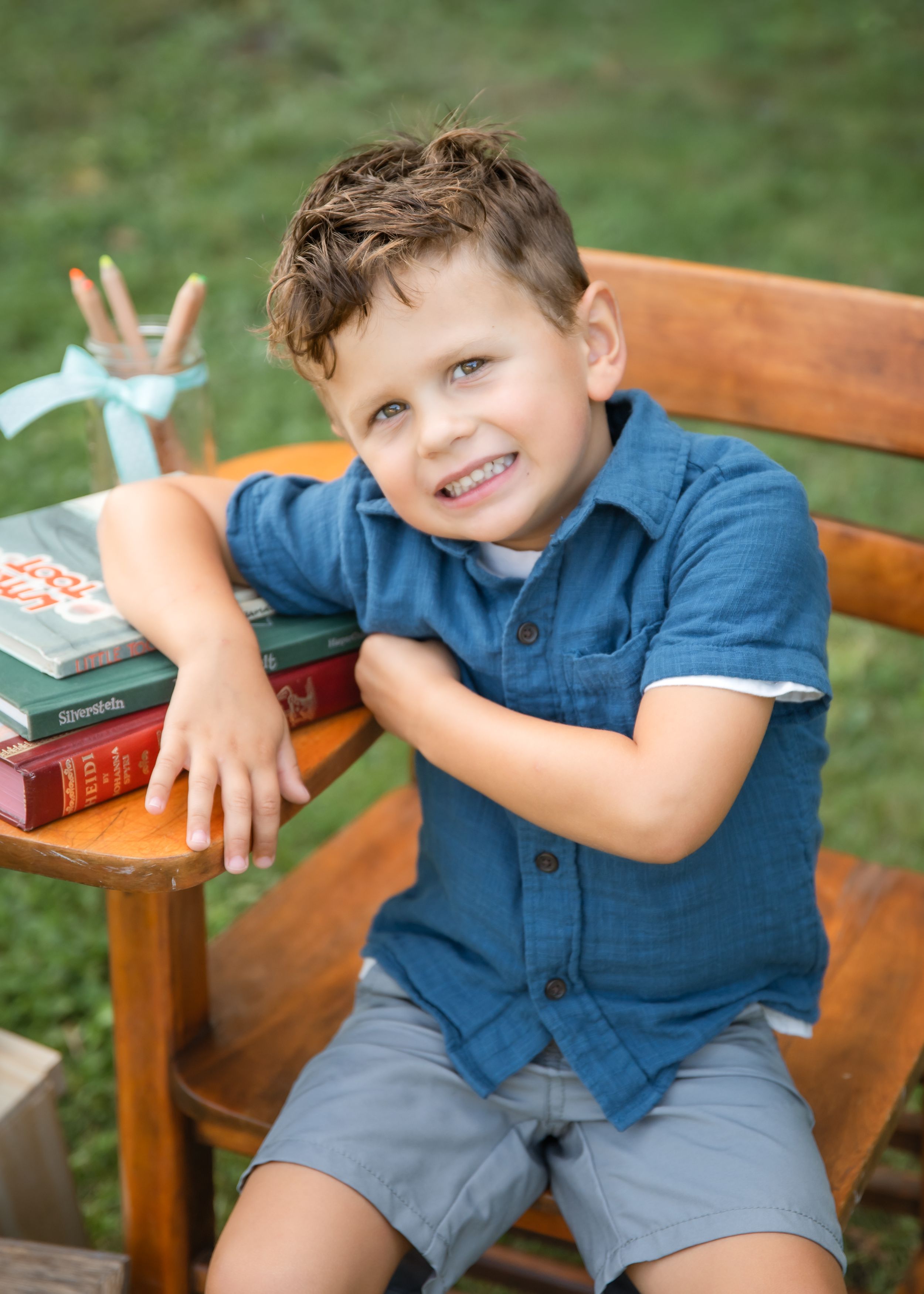 Smiling young boy in blue shirt sitting on wooden bench with stacked books outdoors.