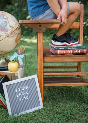 Back to school 4 year Pre-K photo with letter board, globe, apple, books, and child in sneakers on wooden chair.