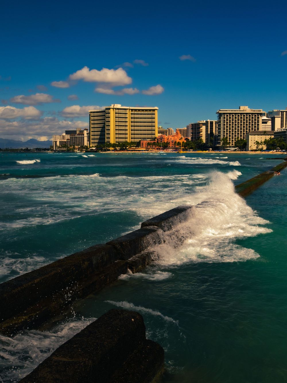Whitewater crashes over a stone breakwater with the pink Royal Hawaiian and Waikiki high-rises under a bright blue sky in Honolulu, Hawaii.