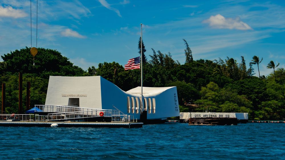 The white USS Arizona Memorial and an American flag rise above blue water at Pearl Harbor, with the USS Arizona BB 39 quay at right.