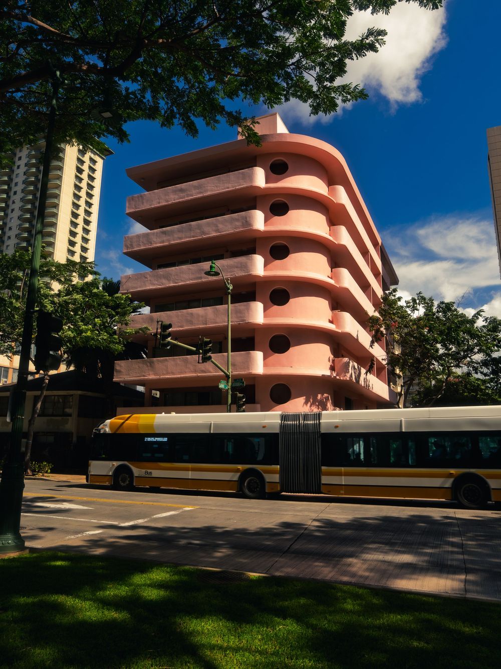 A yellow and white city bus passes a curved pink art deco building on Kuhio Avenue under a blue sky in Waikiki, Honolulu, Hawaii.
