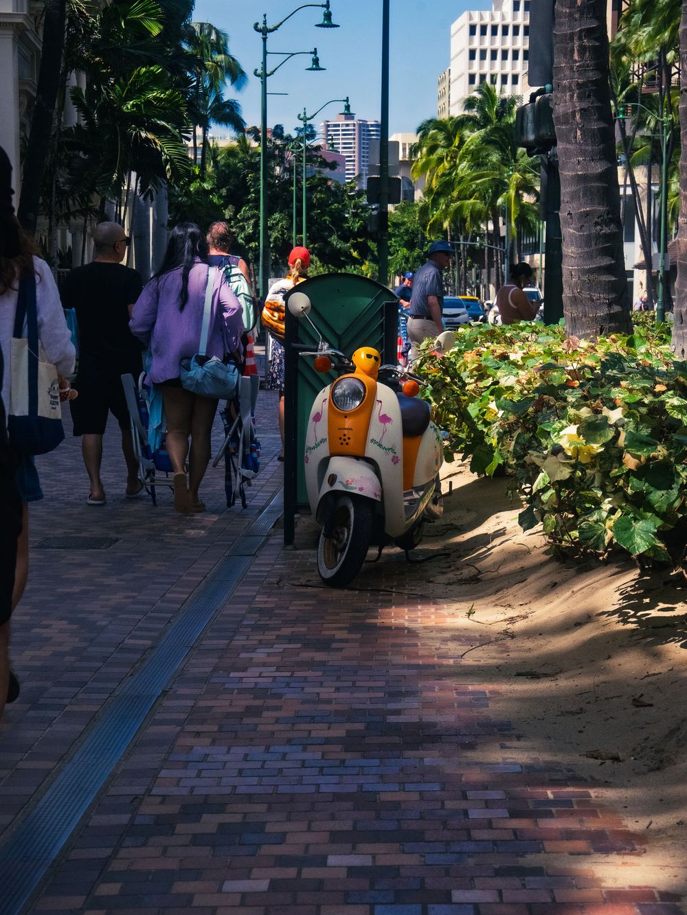 An orange and white flamingo-themed scooter parked on a brick sidewalk as pedestrians stroll past palm trees in Waikiki, Honolulu, Hawaii.