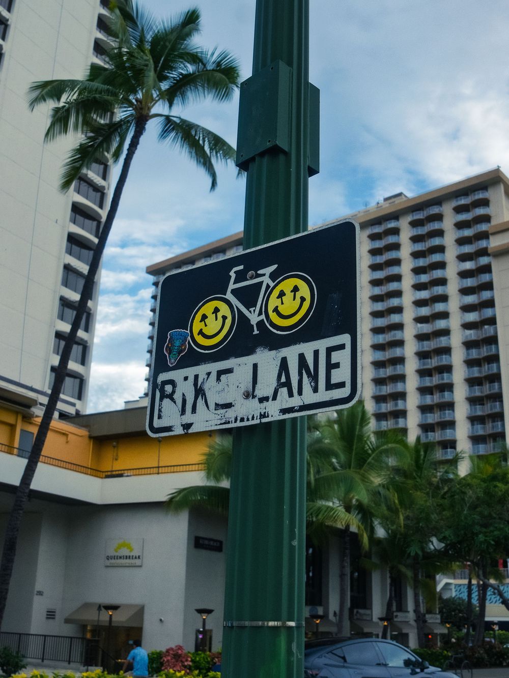 A bike lane sign covered in smiley-face stickers on a green pole, with palm trees and Waikiki hotel towers behind in Honolulu, Hawaii.