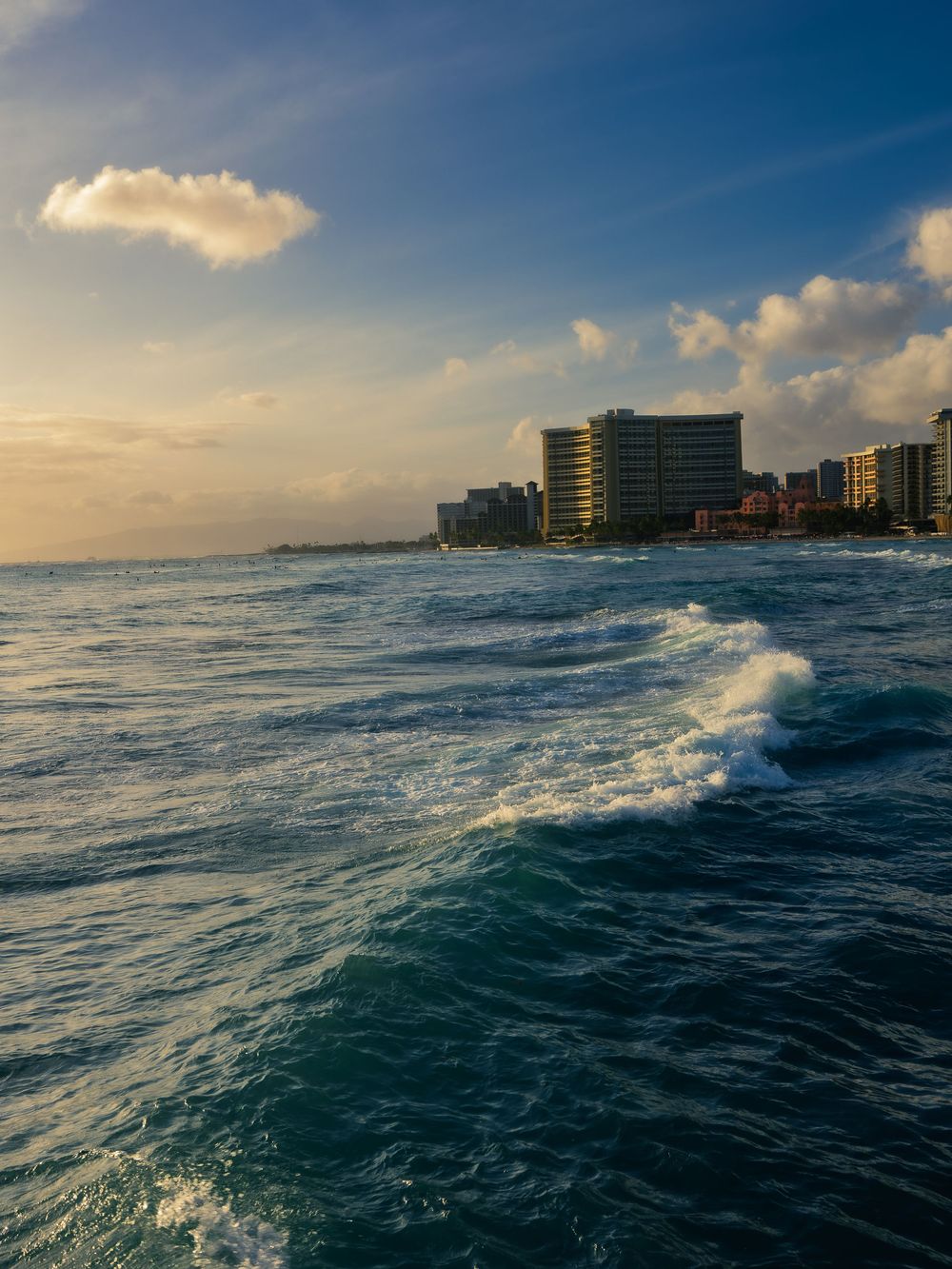 Waves roll toward the Waikiki Beach shoreline in Honolulu at golden hour, with beachfront hotel towers and distant surfers along the coast.