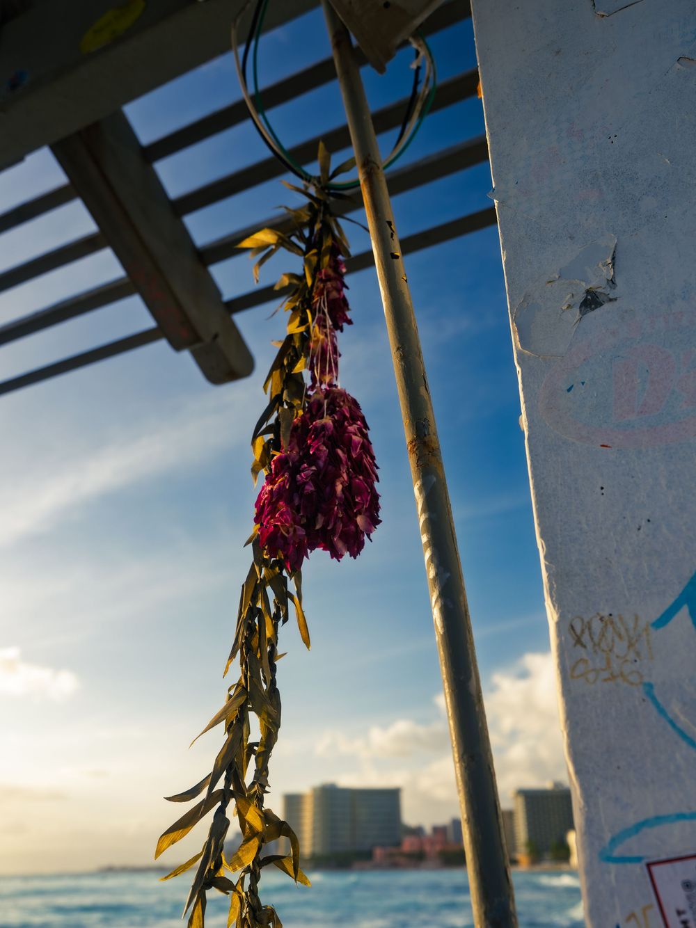 A purple flower lei hangs drying from a weathered structure above the ocean, the Waikiki skyline and blue sky behind in Honolulu, Hawaii.