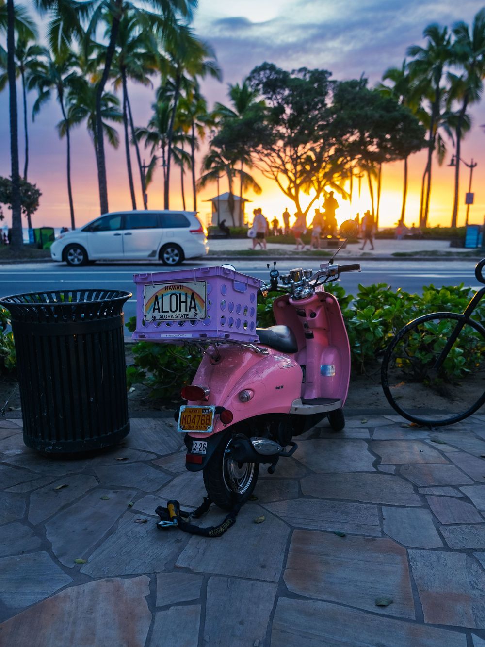 A pink scooter with an ALOHA crate parked on a stone path against a palm-lined sunset over the beach in Waikiki, Honolulu, Hawaii.
