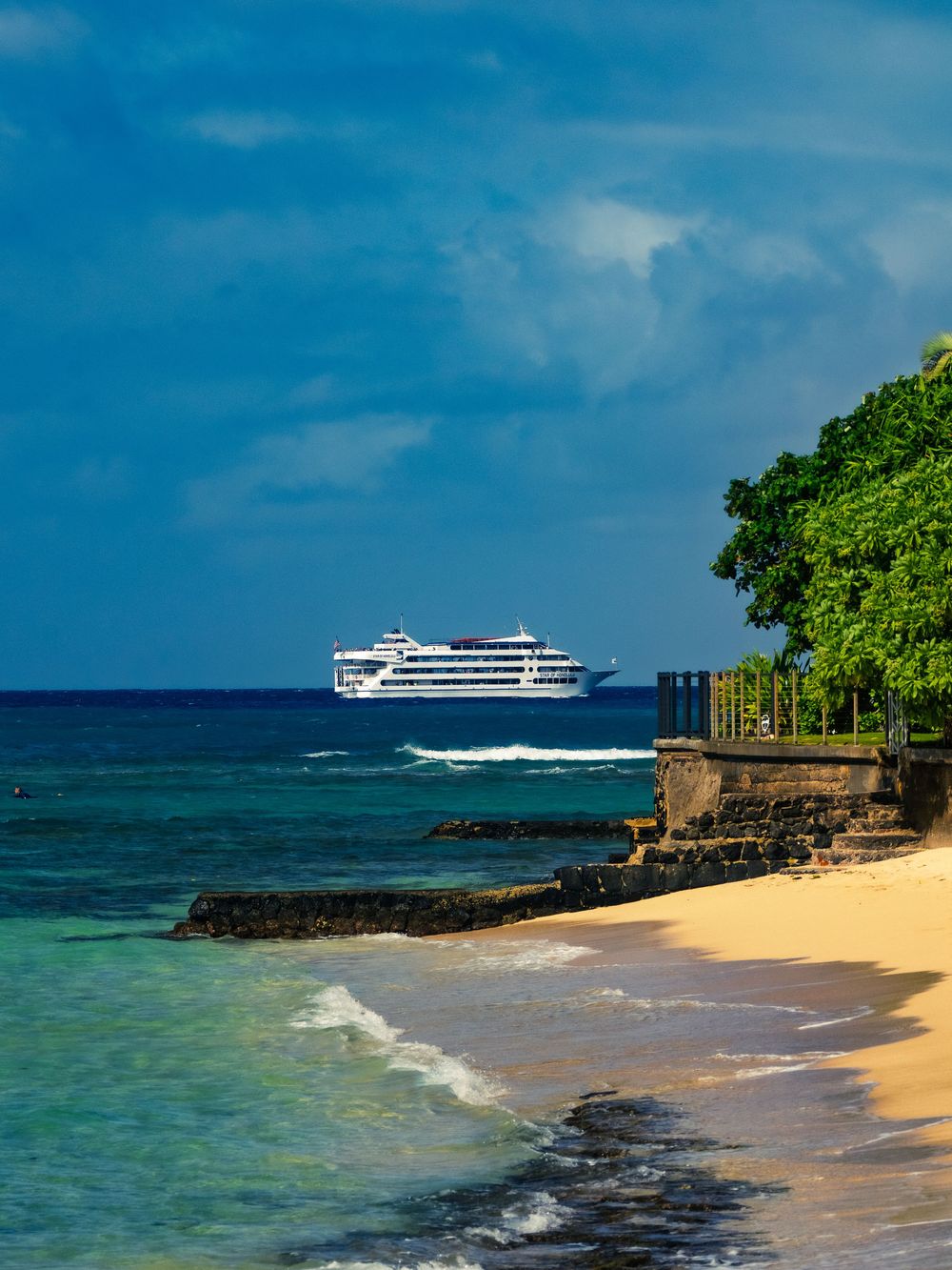 A white cruise boat sails offshore past a golden sand beach and rocky shoreline under a moody blue sky along the coast of Honolulu, Hawaii.