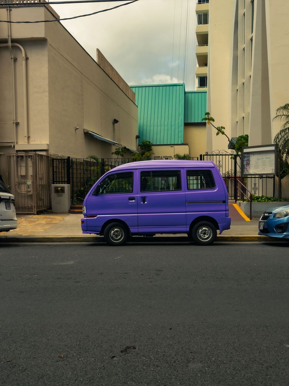 A small purple kei van parked at the curb on a Waikiki street in Honolulu, Hawaii, with beige buildings and a green roof behind it.