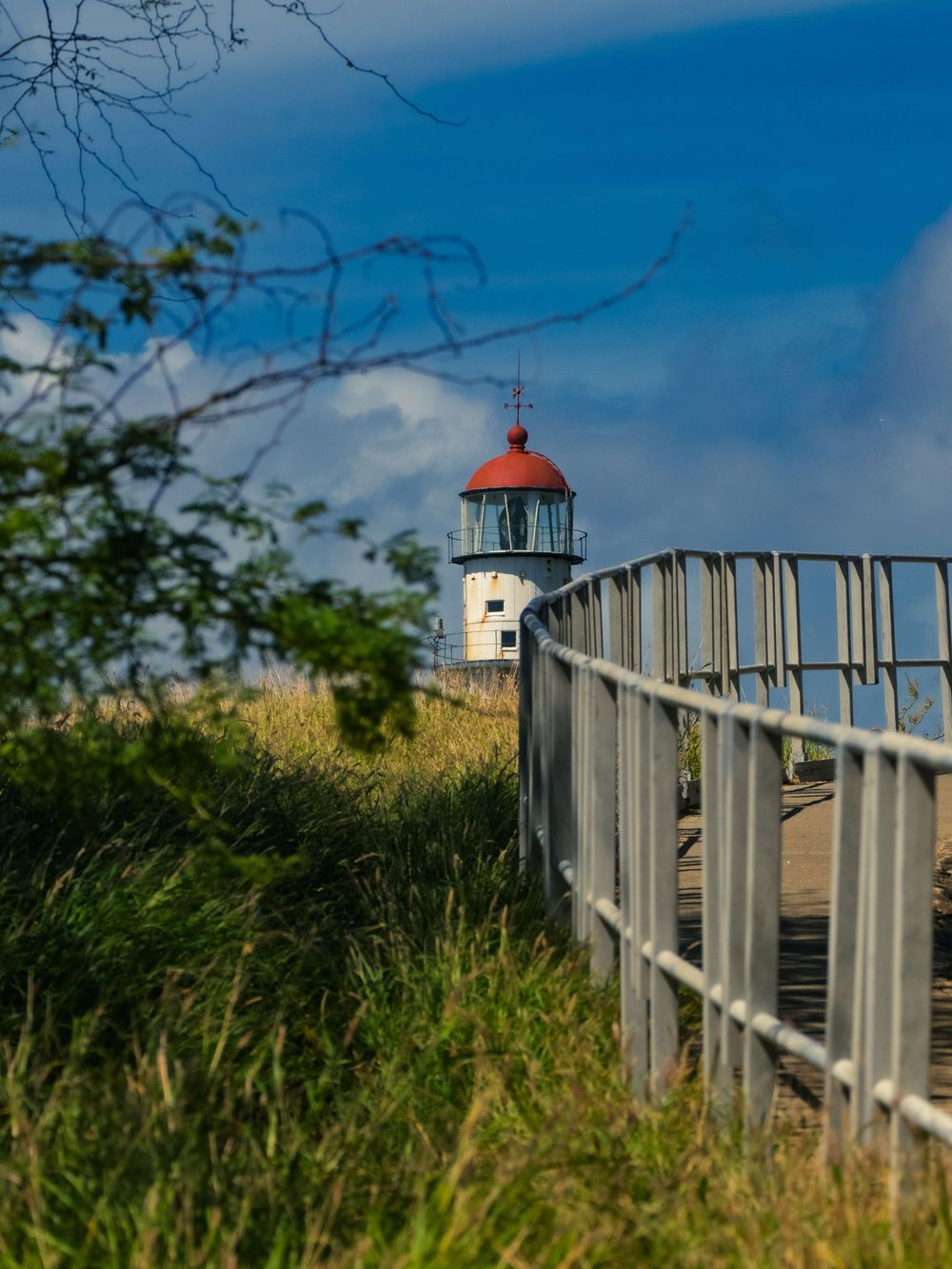 The red-domed Diamond Head Lighthouse sits atop a grassy bluff beyond a curving railed walkway under a blue sky in Honolulu, Hawaii.