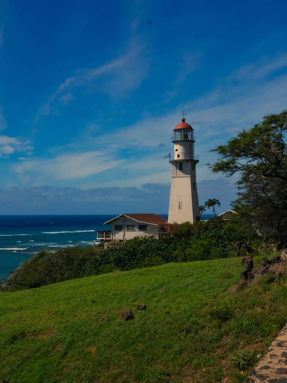 The white, red-domed Diamond Head Lighthouse stands on a green coastal bluff above the blue ocean under a bright sky in Honolulu, Hawaii.