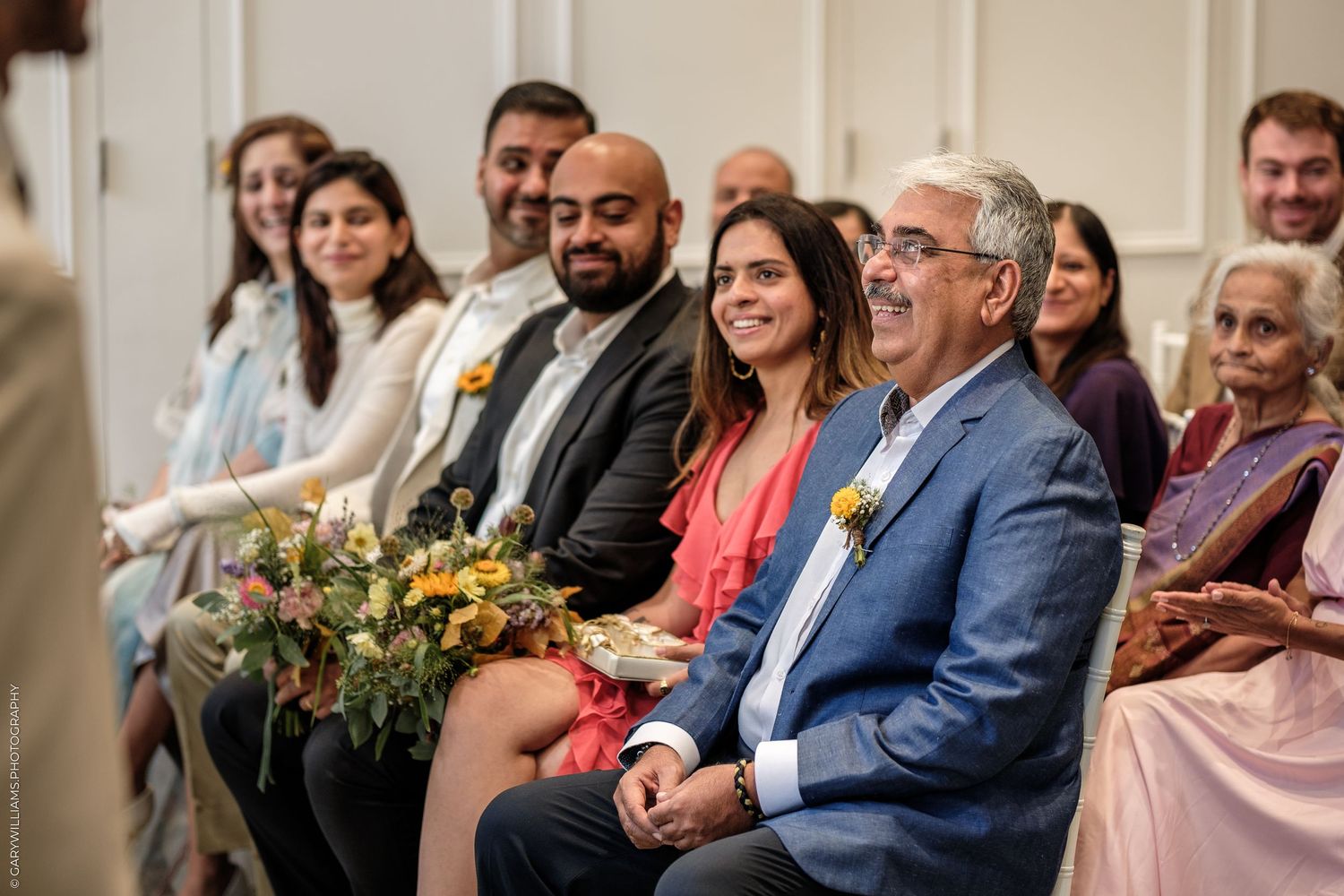 Smiling wedding guests seated in rows at an elegant indoor ceremony, dressed in formal attire with floral arrangements nearby.