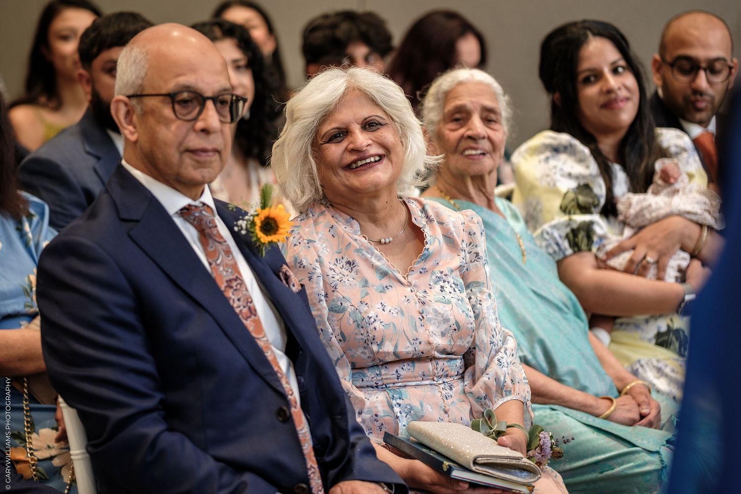 Two smiling adults seated at a formal event, with guests visible in the background.