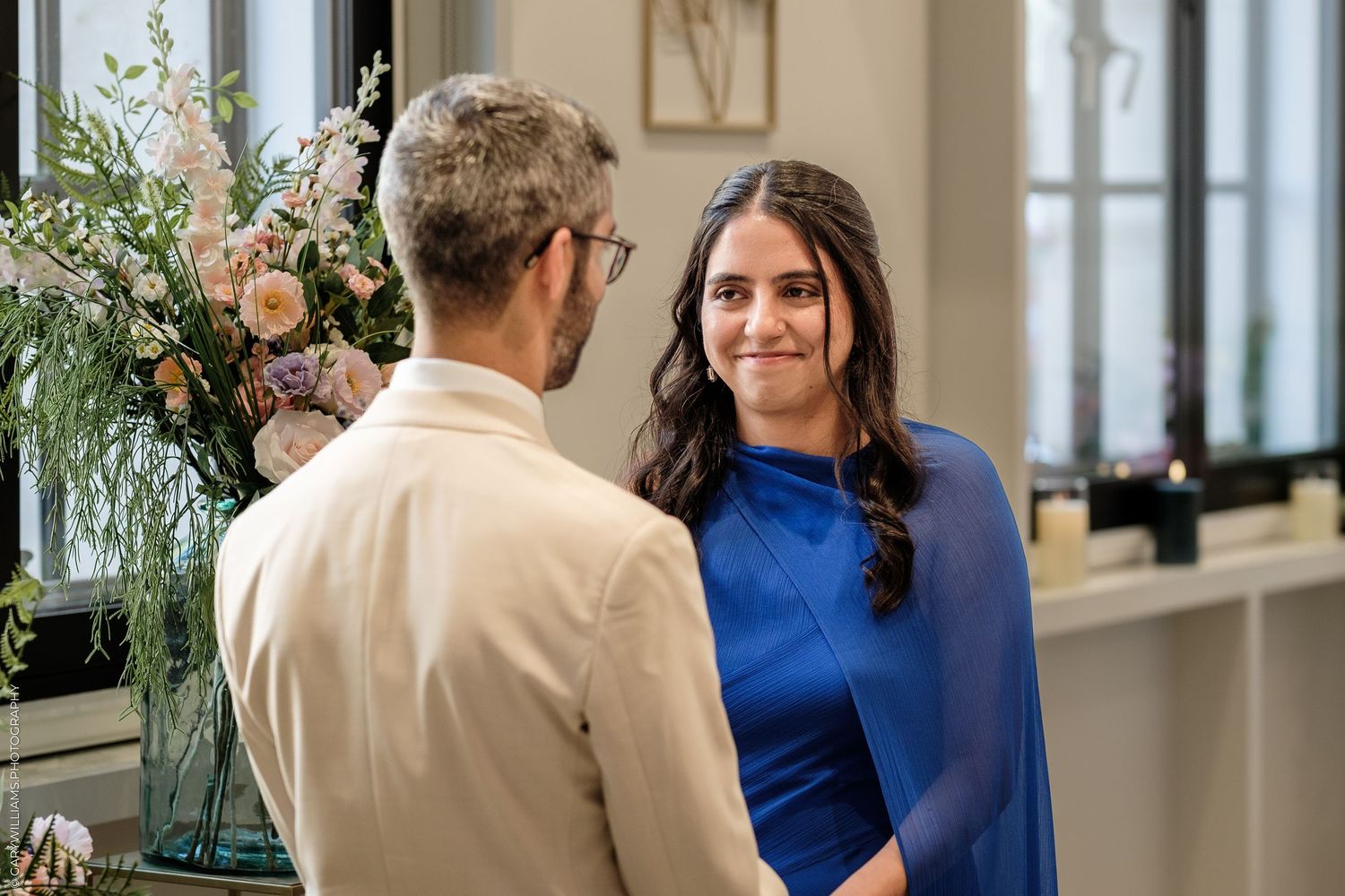 A woman in a blue dress smiles at a man in a beige shirt during an indoor ceremony with floral decor.