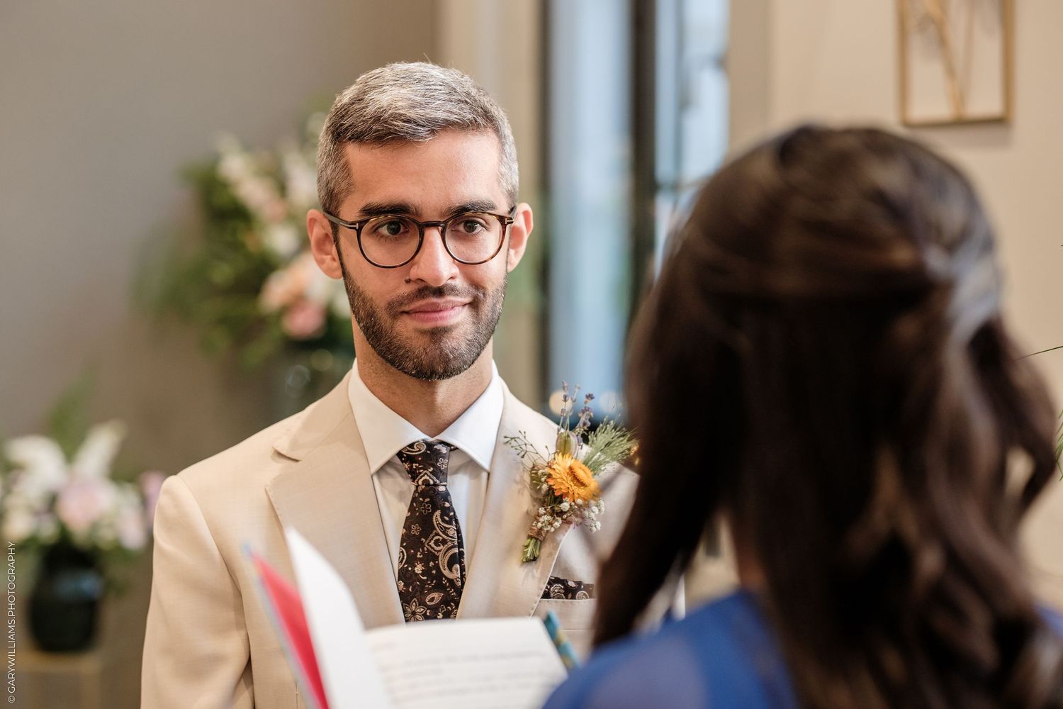 Groom in beige suit with floral boutonniere and patterned tie smiling during wedding ceremony.