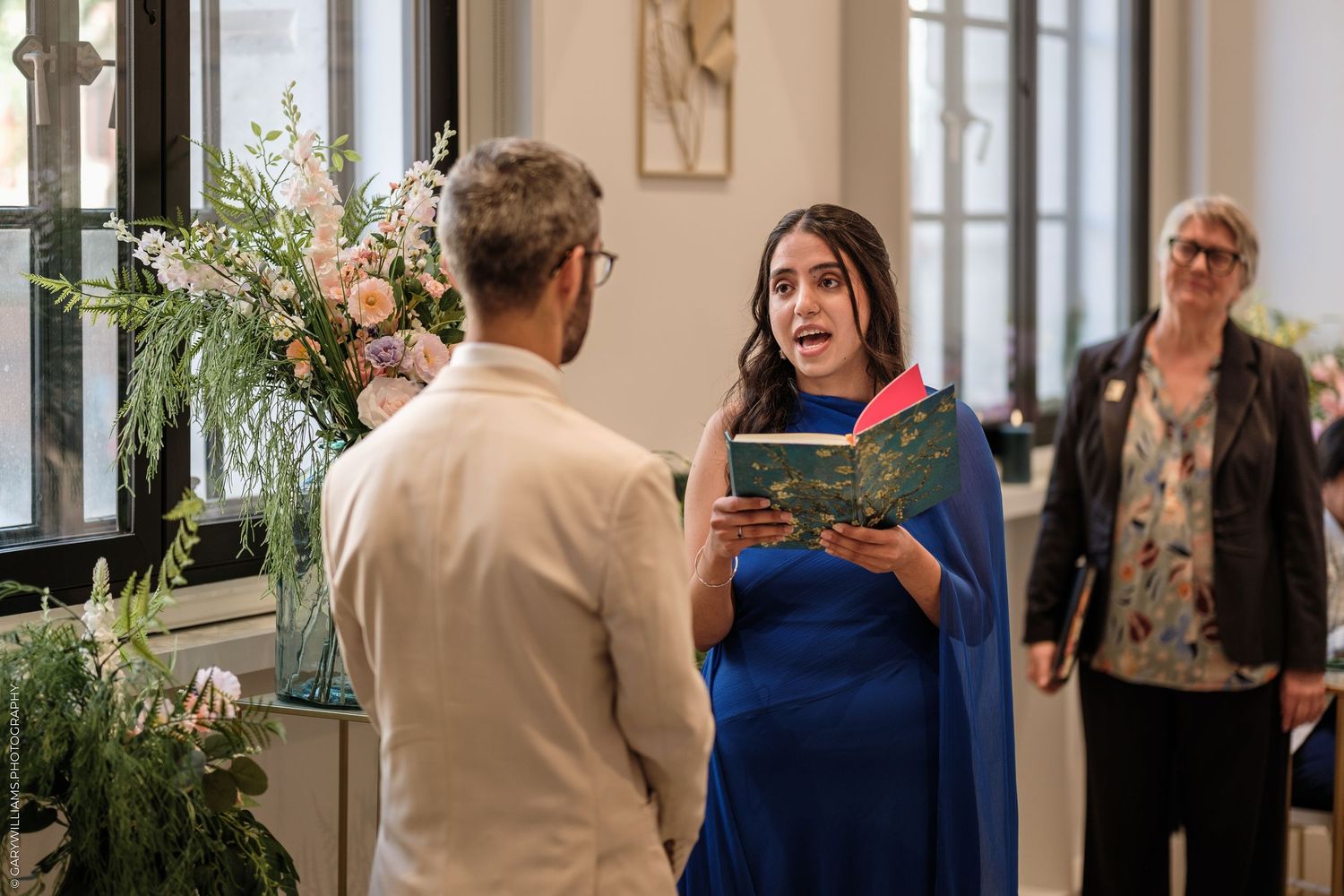 Woman in blue dress reads from book during indoor wedding ceremony with floral arrangement and guests.