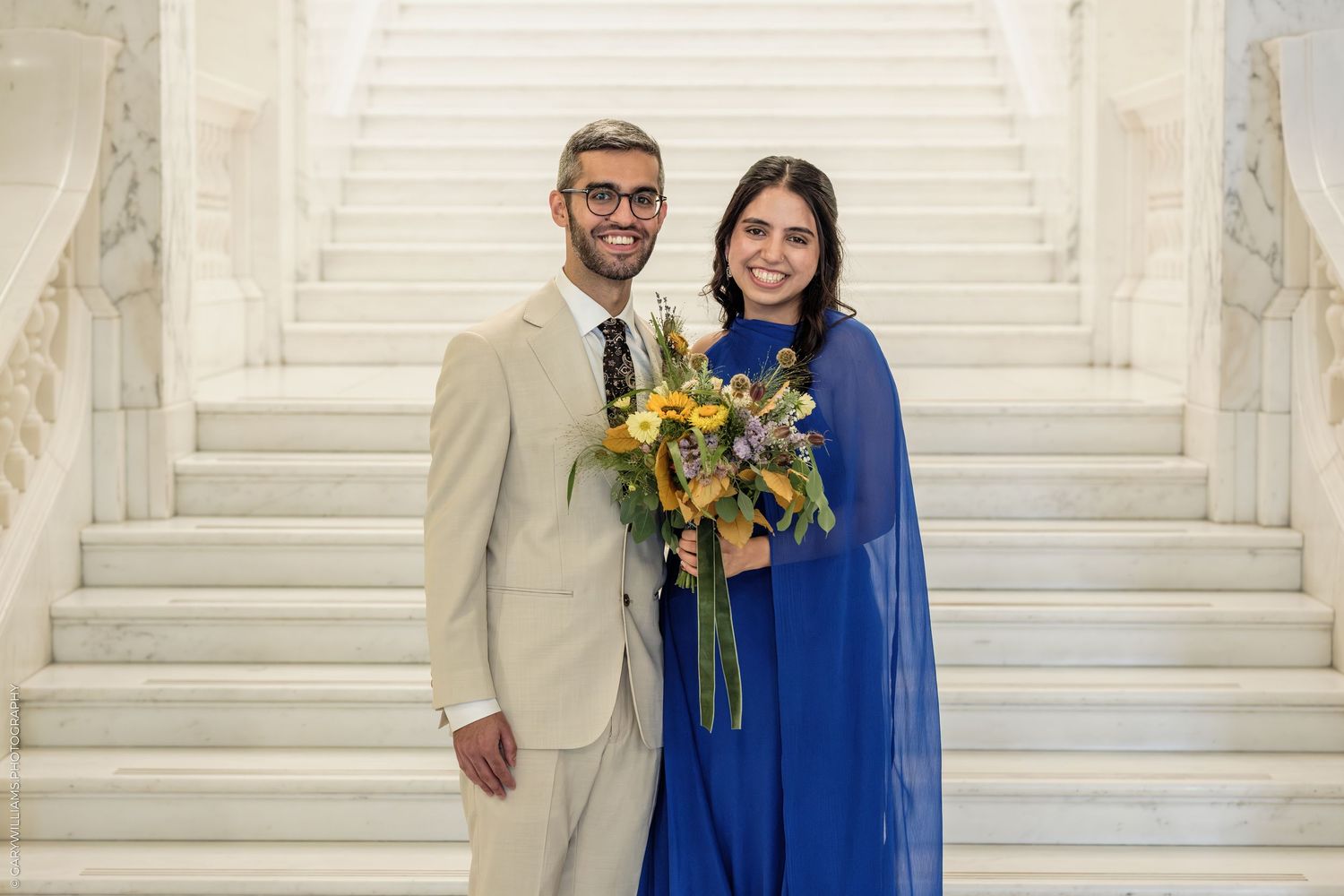 Couple posing on marble staircase, man in beige suit and woman in royal blue dress holding yellow floral bouquet.