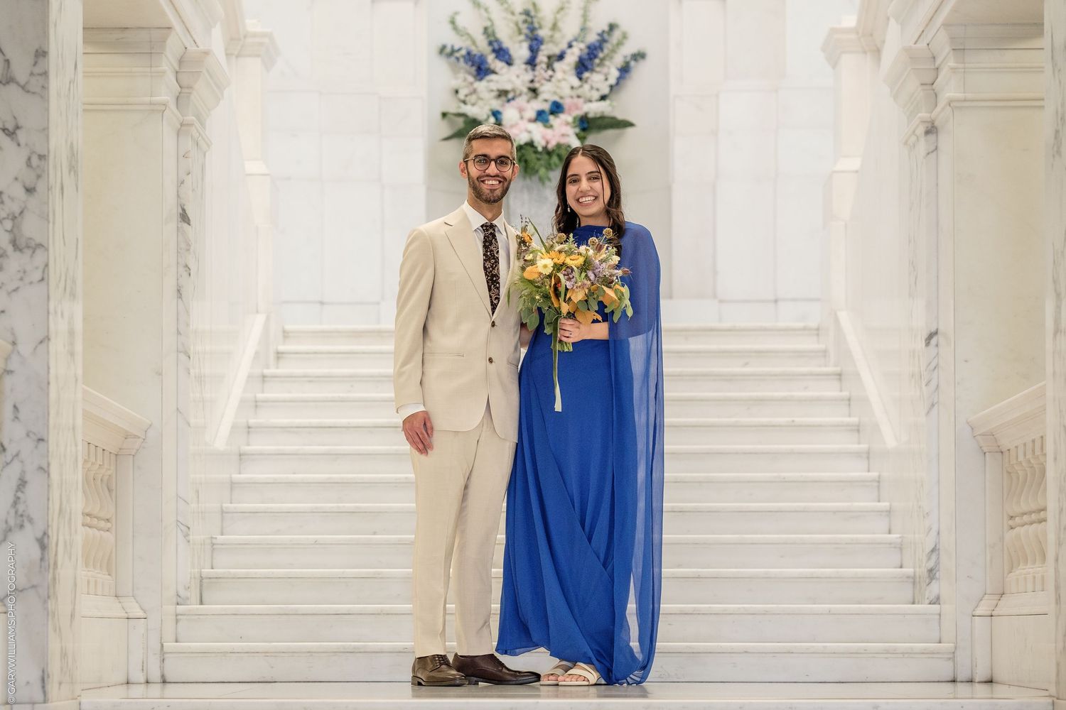 Couple in formal attire, man in beige suit and woman in blue gown, posing on marble staircase with floral bouquet.