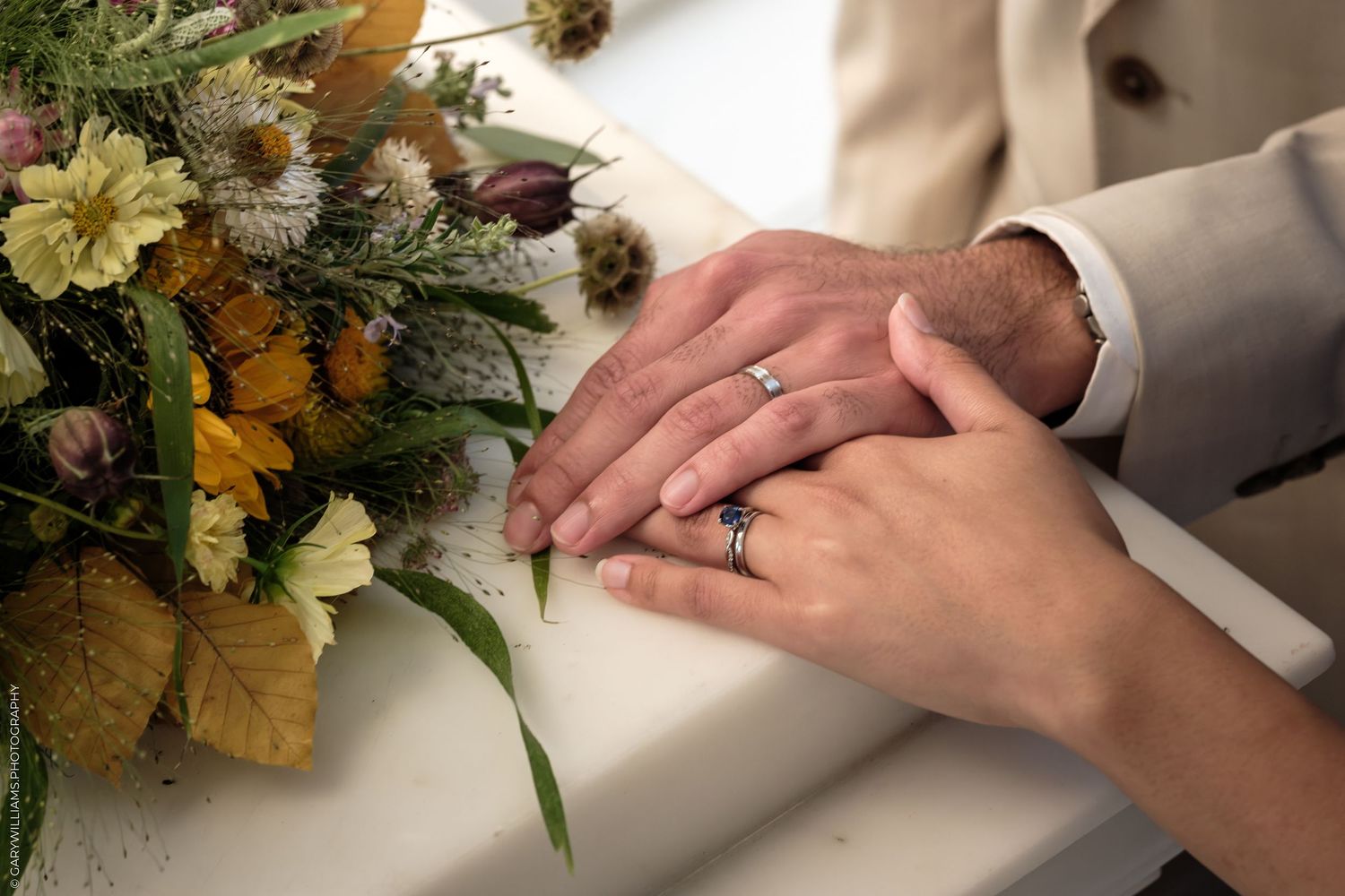 Couple holding hands with wedding rings next to autumn floral bouquet on white surface.