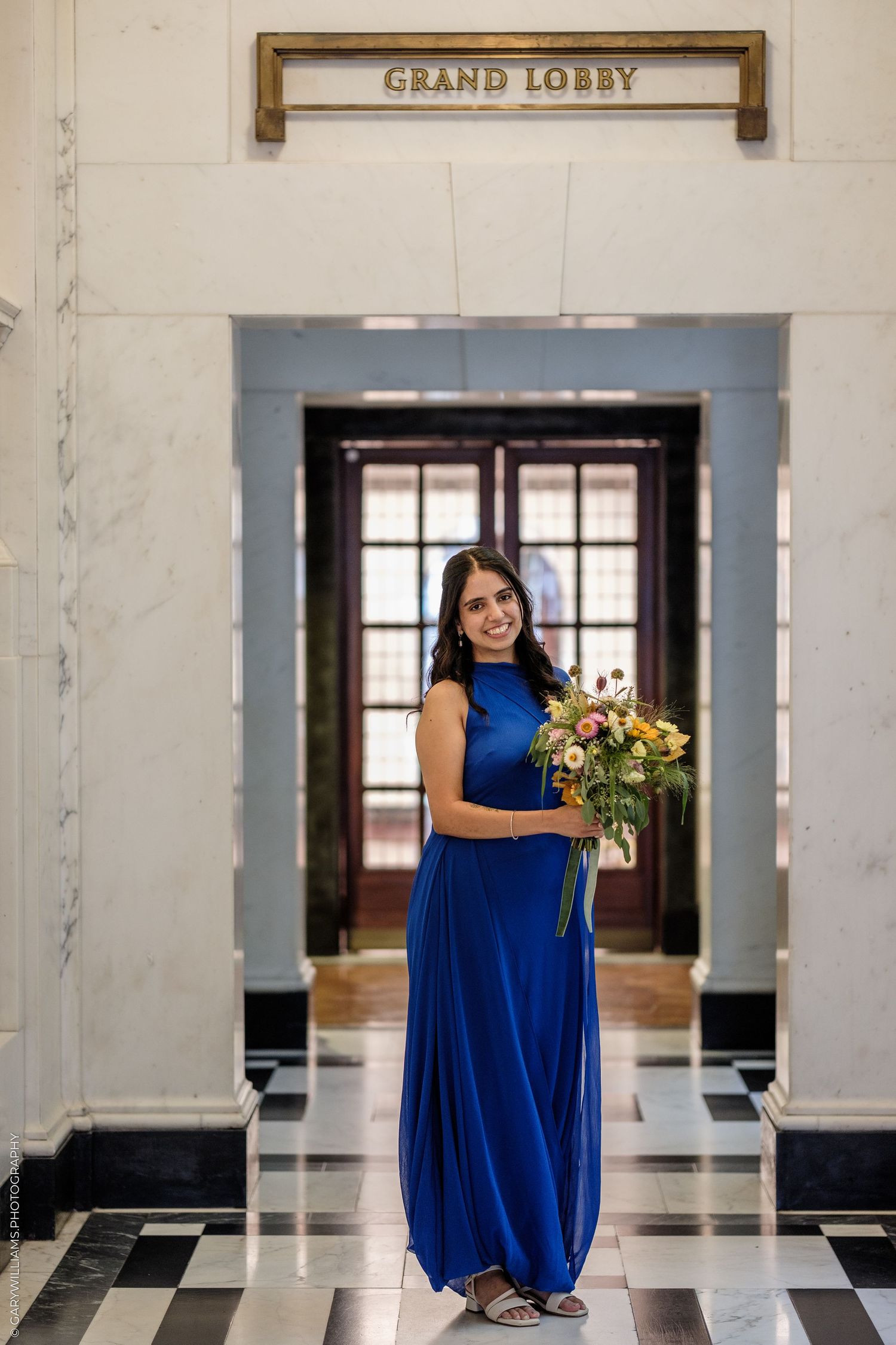 Woman in blue gown holding colorful bouquet standing in Grand Lobby with marble walls and checkered floor.