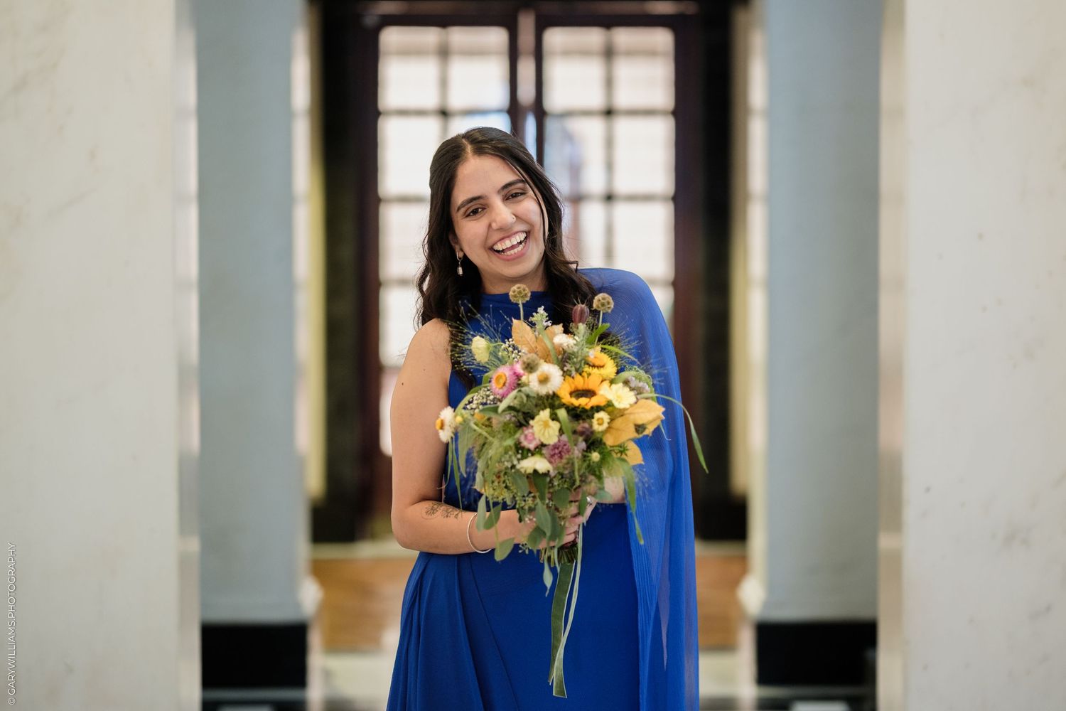 Woman in blue dress smiling and holding colorful wildflower bouquet indoors.