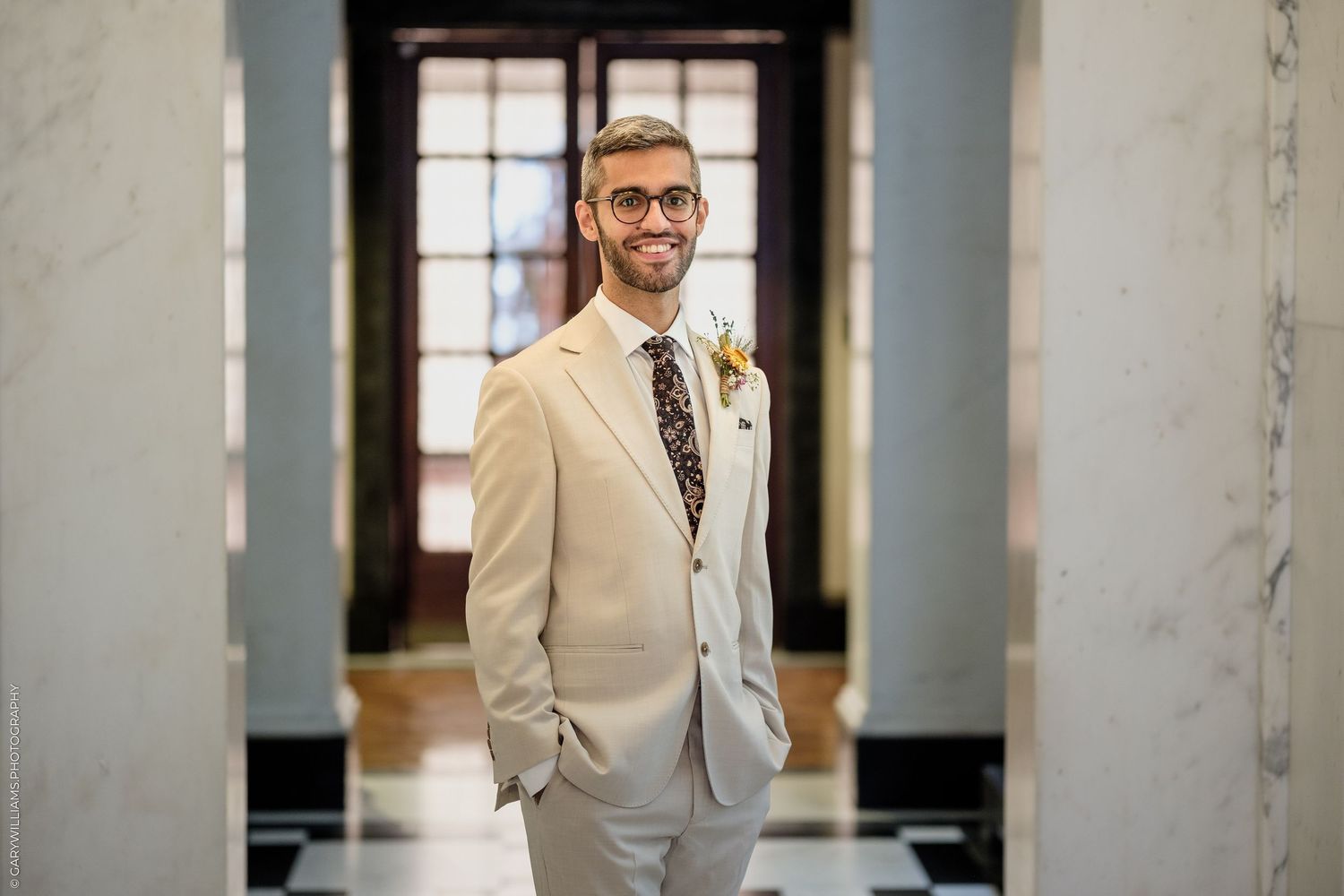Smiling groom in beige suit with floral tie and boutonniere standing in elegant marble hallway.