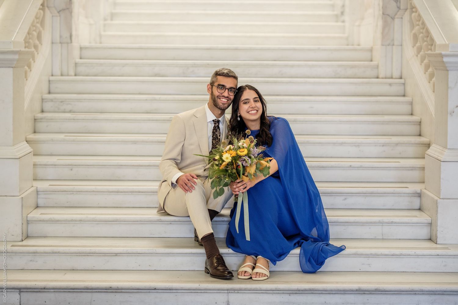 Couple sitting on marble stairs, man in beige suit, woman in blue dress holding yellow floral bouquet, smiling.